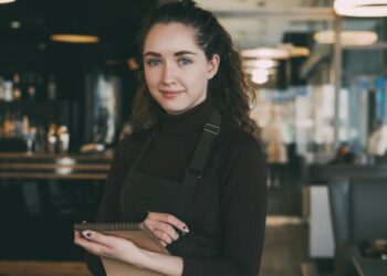 Barista met een zwart schort en coltrui staat in een modern café, houdt een notitieboekje vast en glimlacht naar de camera.
