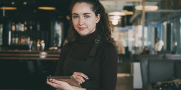 Restaurantmenu's in Singapore in september - GUSTOwebzine Barista met een zwart schort en coltrui staat in een modern café, houdt een notitieboekje vast en glimlacht naar de camera.