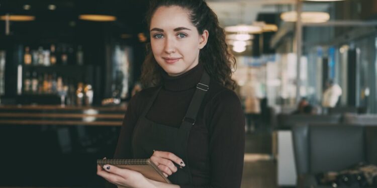 Barista met een zwart schort en coltrui staat in een modern café, houdt een notitieboekje vast en glimlacht naar de camera.