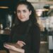 Barista met een zwart schort en coltrui staat in een modern café, houdt een notitieboekje vast en glimlacht naar de camera.