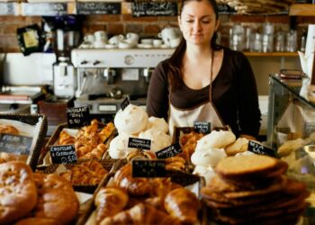 Female bakery worker in a brown shirt and apron stands behind a glass display filled with croissants and pastries.