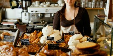 Gluten-free bakeries NYC eat well - GUSTOwebzine Female bakery worker in a brown shirt and apron stands behind a glass display filled with croissants and pastries.
