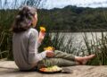 EU regional cheeses at peak in September - GUSTOwebzine Girl sitting barefoot on a wooden dock by a lake, eating orange slices from a metal cup with a plate of food beside her.