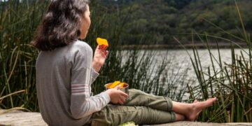 EU regional cheeses at peak in September - GUSTOwebzine Girl sitting barefoot on a wooden dock by a lake, eating orange slices from a metal cup with a plate of food beside her.