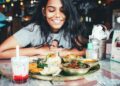Eine junge Frau lächelt, während sie an einem Tisch mit einer großen indischen Mahlzeit im Thali-Stil und einem Glas Getränk in einem Restaurant sitzt.