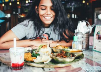 Young woman smiles while seated at a table with a large thali-style Indian meal and a glass of beverage in a restaurant setting.