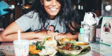 Eine junge Frau lächelt, während sie an einem Tisch mit einer großen indischen Mahlzeit im Thali-Stil und einem Glas Getränk in einem Restaurant sitzt.