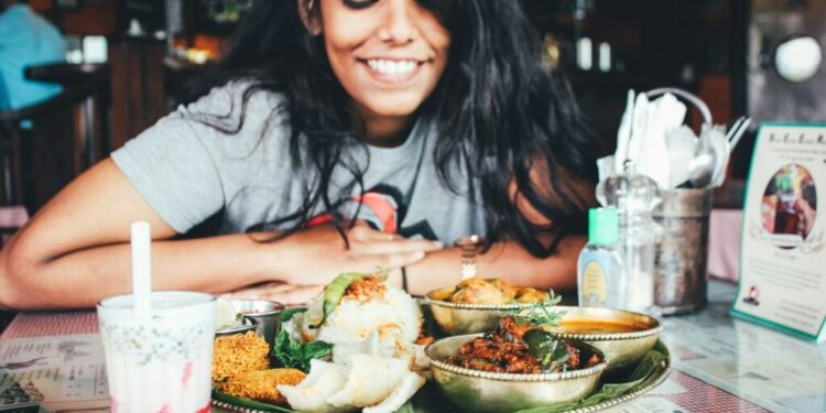 Eine junge Frau lächelt, während sie an einem Tisch mit einer großen indischen Mahlzeit im Thali-Stil und einem Glas Getränk in einem Restaurant sitzt.