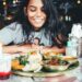 Young woman smiles while seated at a table with a large thali-style Indian meal and a glass of beverage in a restaurant setting.