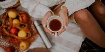 vrouw-met-kop-gekruide-thee-herfst-unsplash - GUSTOwebzine Een vrouw houdt een keramieken mok met warme gekruide dranken vast naast herfstbladeren, wat het seizoensgebonden comfort van dranktradities laat zien.