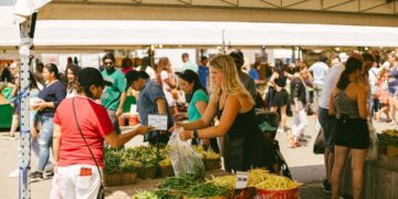 vrouw-shopt-op-boerenmarkt-unsplash - GUSTOwebzine Een vrouw kiest seizoensgroenten op oogstmarkten in Europa tijdens september, met kratten vol verse producten uitgestald.