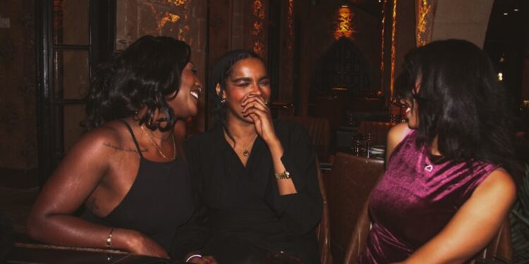 Three women sit together at a dimly lit restaurant table, laughing and chatting warmly while wearing stylish outfits.