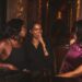 Three women sit together at a dimly lit restaurant table, laughing and chatting warmly while wearing stylish outfits.