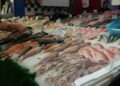 Seafood stall with multiple fish laid on crushed ice and handwritten price signs at a market.