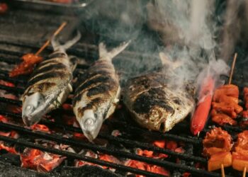 Two whole grilled fish with charred skin on a barbecue grill, with skewered red sausages and meat chunks beside them.