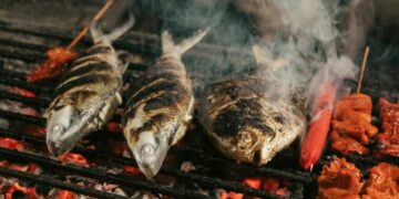 Two whole grilled fish with charred skin on a barbecue grill, with skewered red sausages and meat chunks beside them.