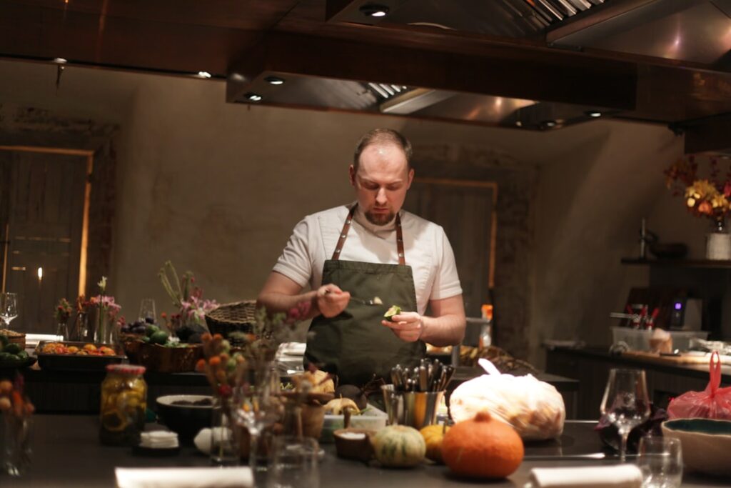 Chef con camisa blanca y delantal verde sirve la comida en una concurrida barra de cocina, rodeado de verduras y cuencos.