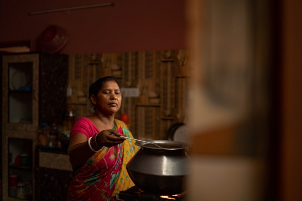 Une femme indienne en sari coloré cuisinant sur un pot en métal sur un poêle, tenant une louche dans une cuisine faiblement éclairée.