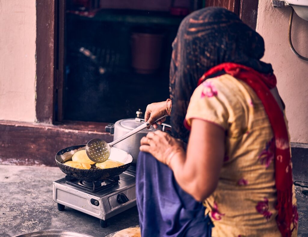 Una donna in un abito colorato e sciarpa rossa friggere pani rotondi su una padella sopra un fornello a gas portatile fuori da un'entrata.