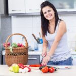 Uma mulher organizando cuidadosamente recipientes de receitas de piquenique em uma cesta de vime na cozinha, pronta para um passeio ao ar livre em junho.