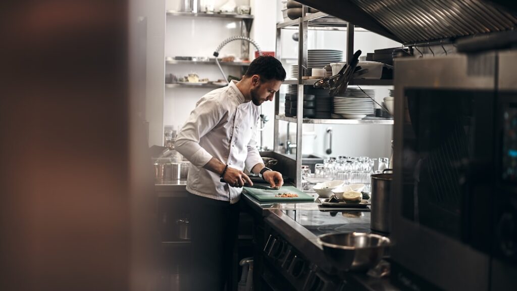 Chef en veste blanche hachant des ingrédients sur une planche à découper verte à un comptoir de préparation en acier inoxydable dans une cuisine animée