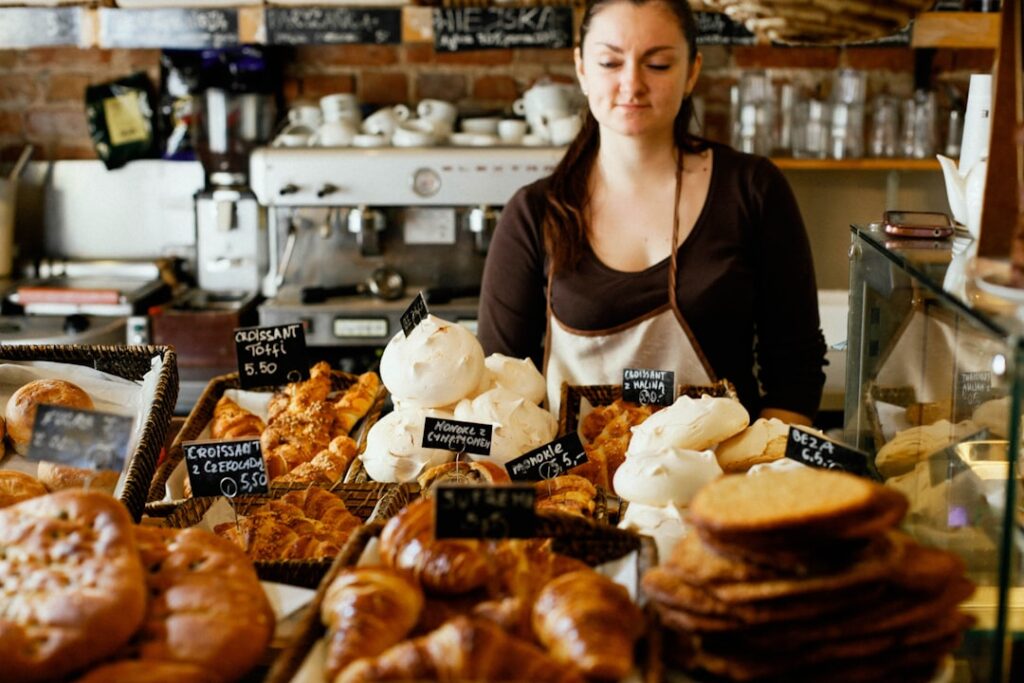 Una trabajadora de panadería se sitúa detrás de una vitrina de cristal llena de croissants y pasteles en una cafetería.