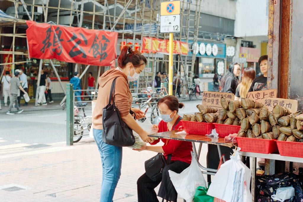 Woman in a mask buys sticky rice dumplings from a street vendor at an outdoor market stall with red baskets and stacked wraps on display.",