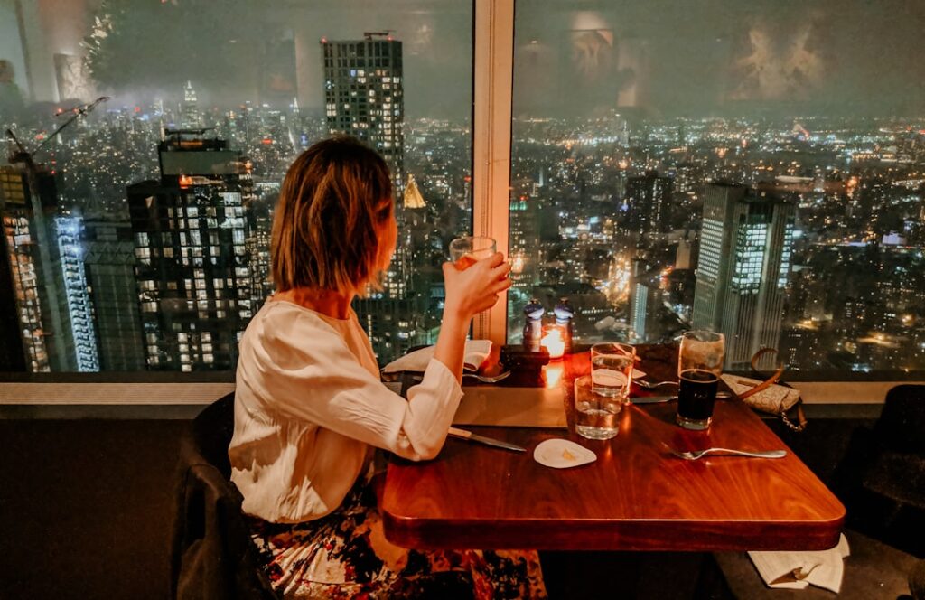 Femme assise à une table de restaurant à la lumière des bougies, trinquant avec un verre tandis que les lumières de la ville brillent par la fenêtre derrière elle la nuit.