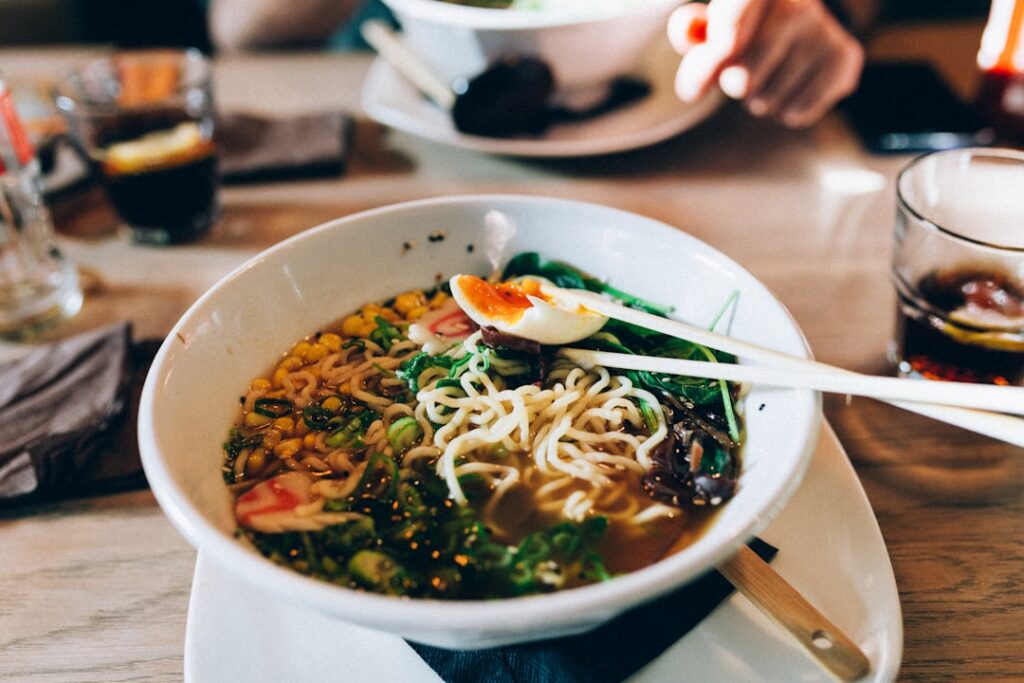 Un bol de ramen avec des nouilles, du maïs, des légumes verts et un œuf à moitié cuit, des baguettes posées dessus sur une table en bois dans un cadre de salle à manger.