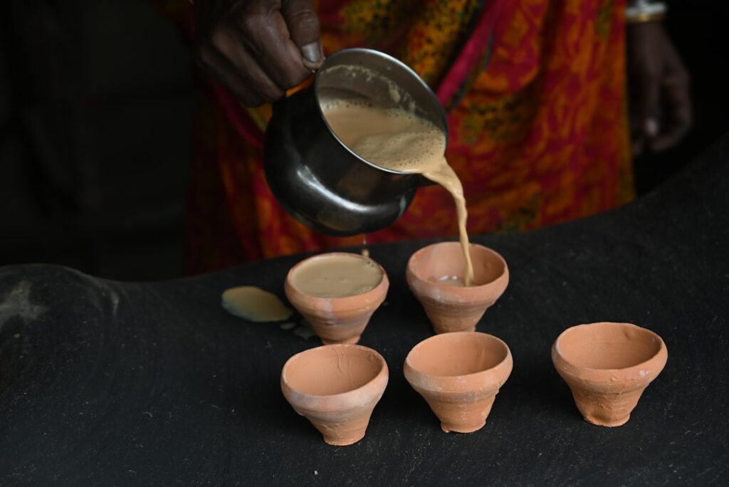 Person pours beige batter from a metal pitcher into five small terracotta cups on a dark surface.
