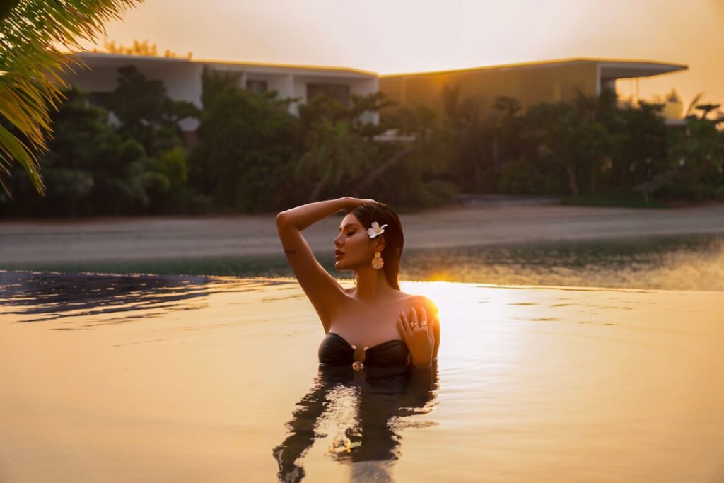Donna in bikini nero in piedi in una piscina illuminata dal tramonto fino al petto, con la mano appoggiata sulla testa e un fiore tra i capelli.