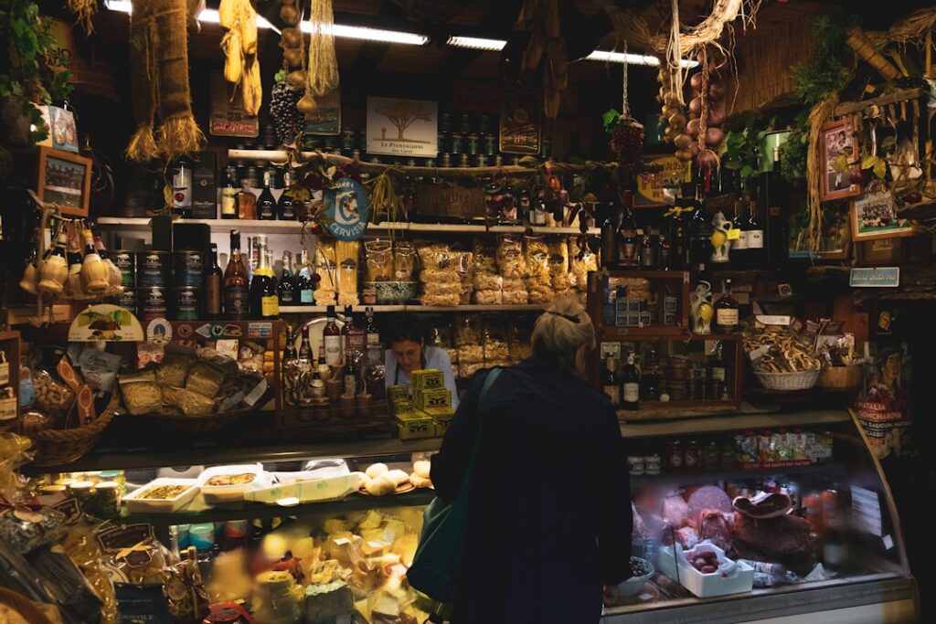 Puesto de mercado acogedor con estantes de botellas, tarros y alimentos secos; un cliente explora la pantalla del mostrador detrás del cristal.