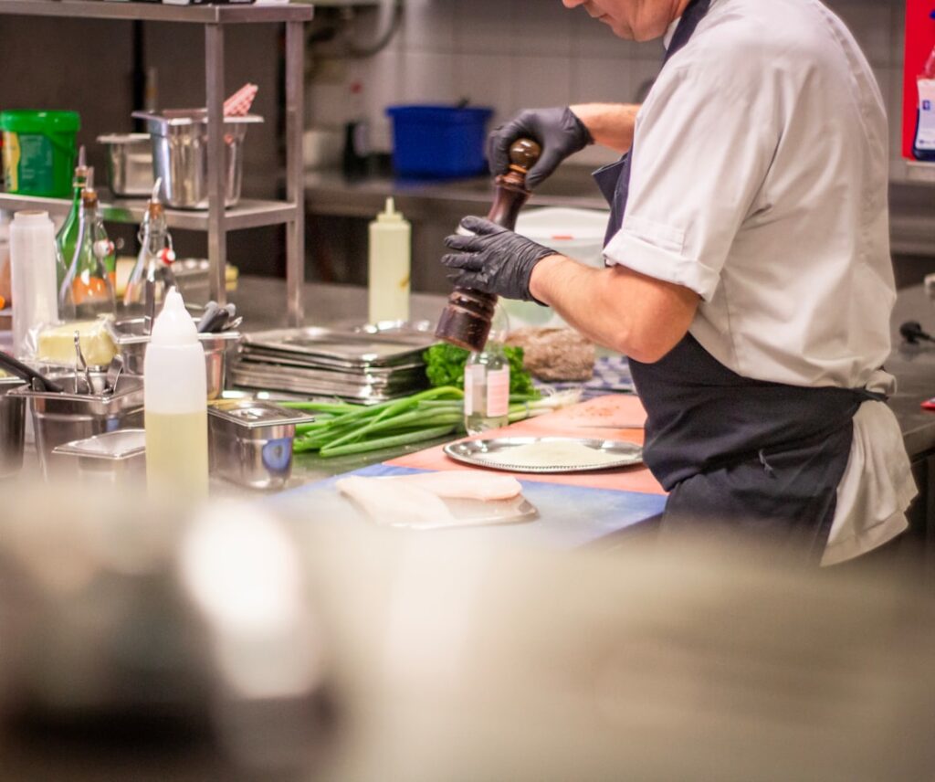 Chef in a white uniform and gloves grinds pepper onto meat at a busy kitchen prep station, with greens and utensils around.
