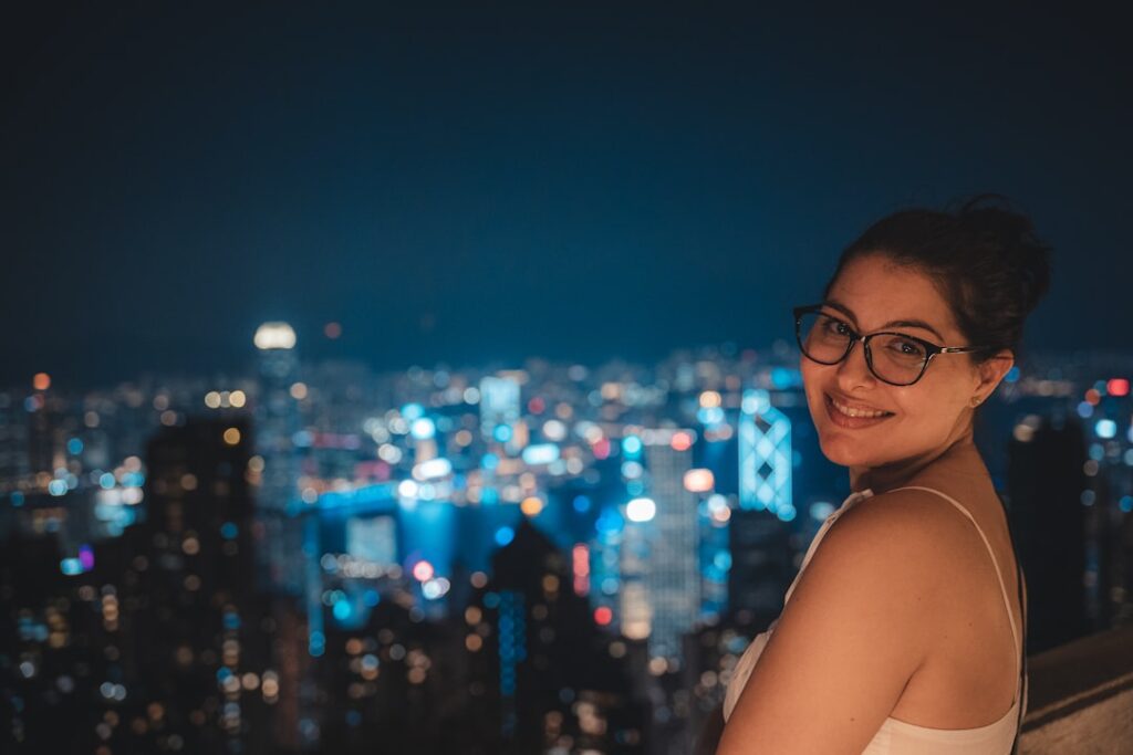 Smiling woman with glasses on a rooftop at night, city skyline blurred in the background.
