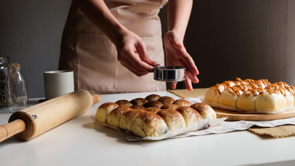Baker presses a round metal cookie cutter into dough balls on a white countertop, with a rolling pin and two trays of bread buns nearby.