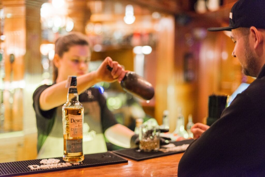 Barman a servir uísque de uma garrafa para um shaker para um cliente num bar ao balcão.