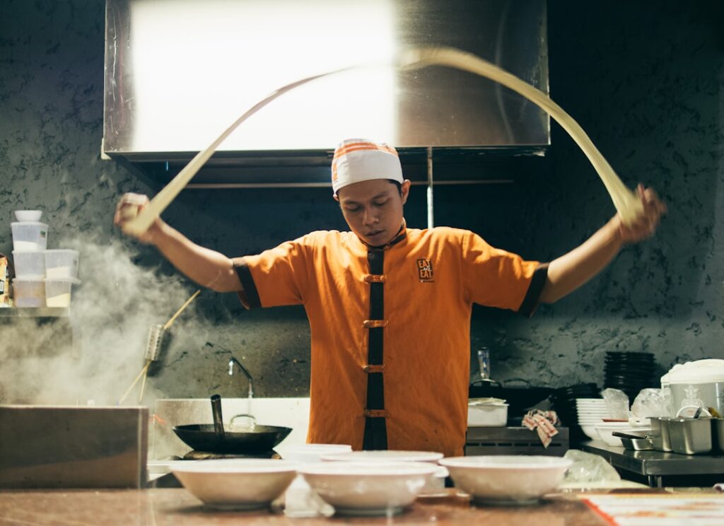 Chef in orange uniform tossing dough in a busy kitchen with steam and bowls on the counter behind him