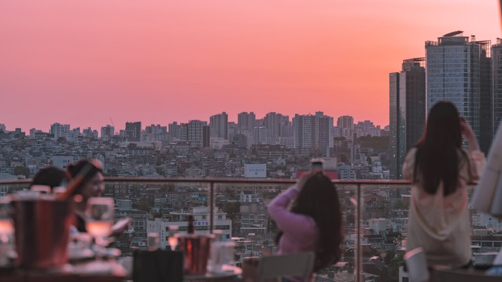 People dining on a rooftop at sunset with a pink-orange sky over a city skyline.