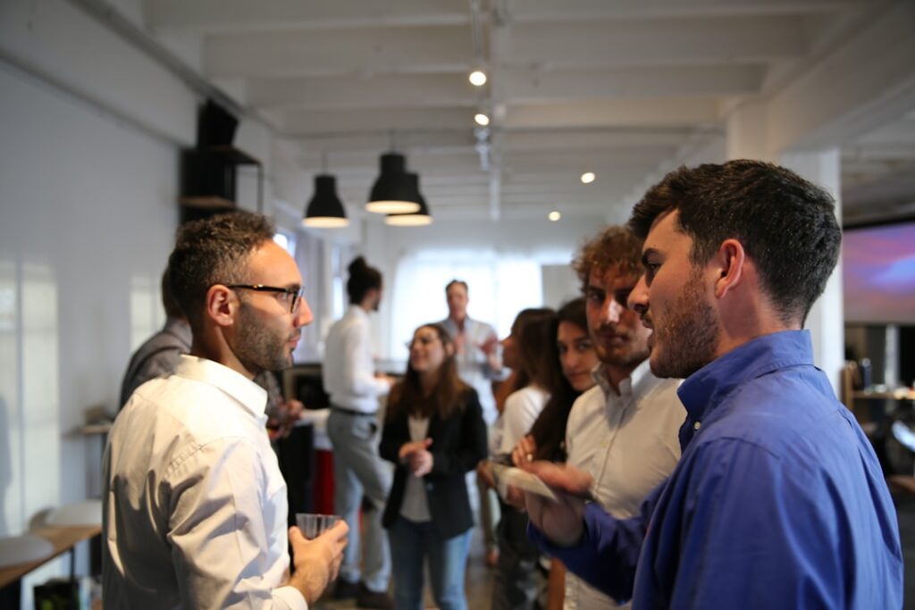 Group of coworkers networking at an office party, some holding drinks and chatting in a bright open space with others in the background.]