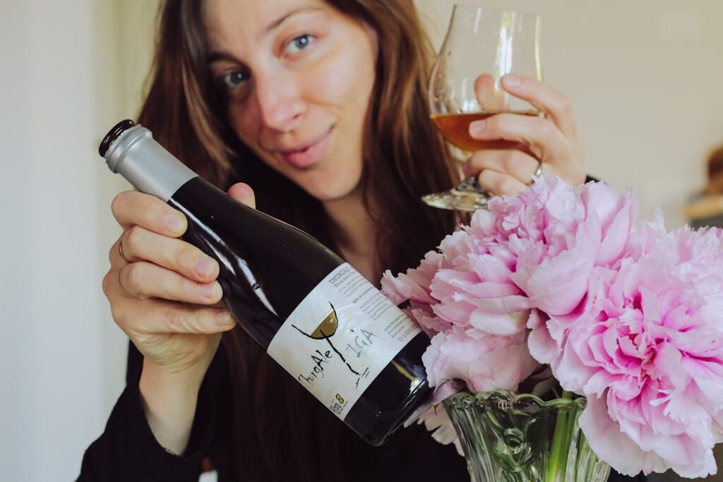 Woman holding a wine bottle and glass, smiling, with pink flowers in a vase nearby as she pours wine.