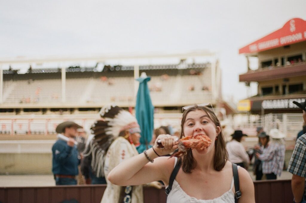 Young woman in a white top bites into a chicken wing at an outdoor fair, with a man in a feather headdress and other attendees in the background.