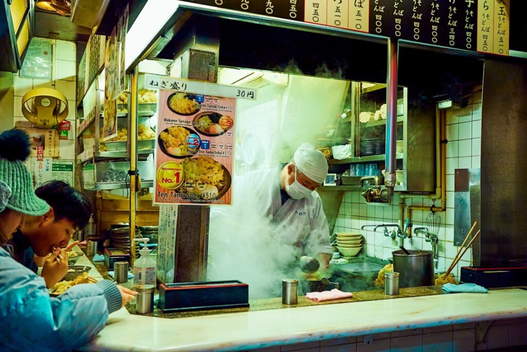 Balcão de loja de macarrão japonês aberto com um chef de chapéu branco e máscara cozinhando em meio a vapor subindo, enquanto clientes comem no bar à esquerda.