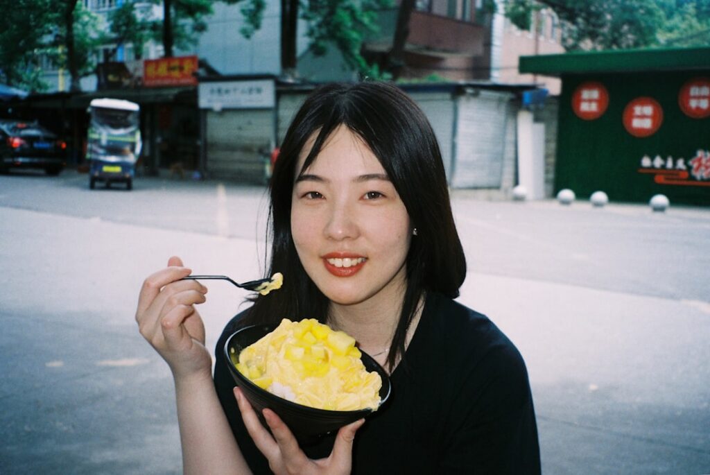 Young woman outdoors, smiling while eating a bowl of mango shaved ice with a spoon.