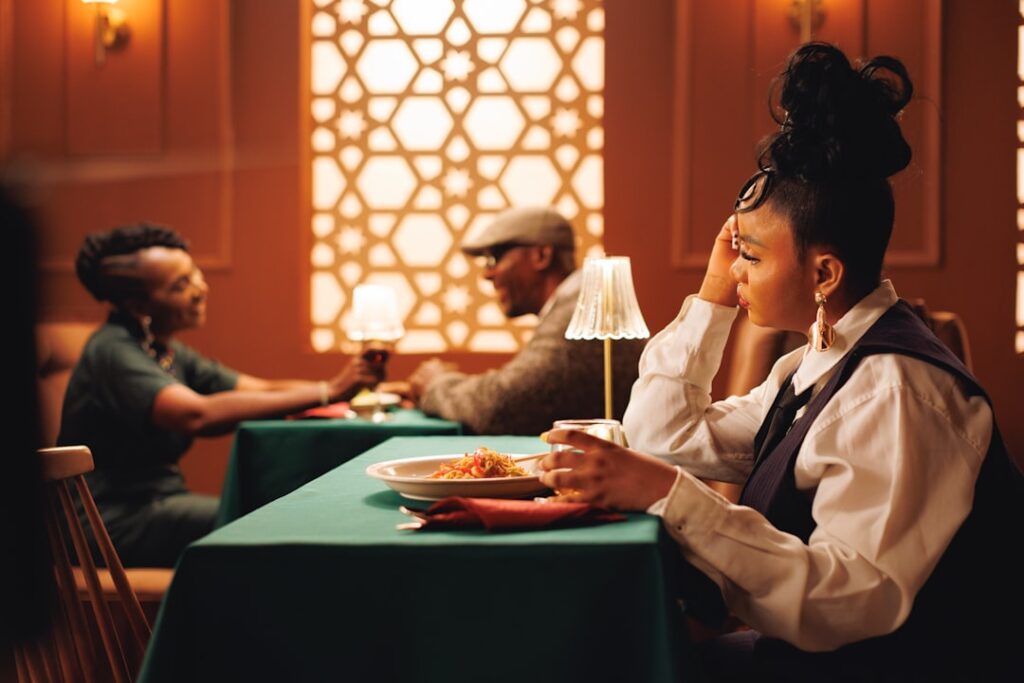 Woman with an elaborate bun sits at a green-clothed table in a warmly lit restaurant, resting her head on her hand as she looks away; plate of pasta in front.