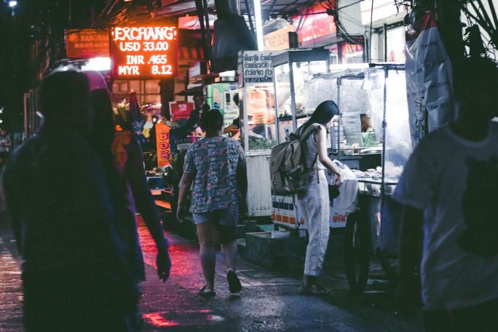 Night market street at night with neon signs; a woman with a backpack buys food from a stall as other pedestrians walk by, wet pavement reflecting lights.