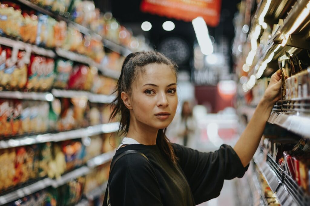 Donna che fa shopping in un corridoio di supermercato, allungando la mano per afferrare un articolo dallo scaffale.