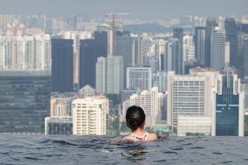Back view of a person with dark hair in an infinity pool, facing a dense city skyline.
