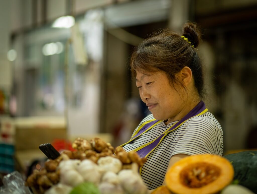 Venditrice di mercato, una donna in camicia a righe che guarda il telefono mentre sta in piedi vicino a mucchi di prodotti tra cui aglio e zenzero in un banco di mercato affollato.