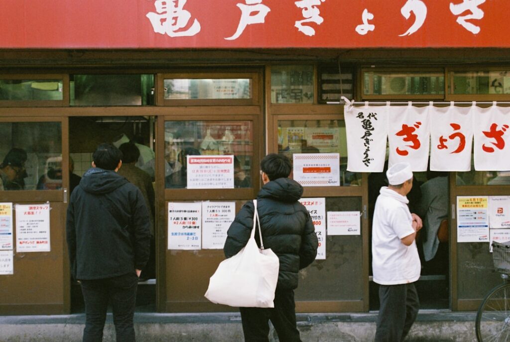People stand in line outside a Japanese eatery with white curtain banners and red signage above the door, posters on the windows.