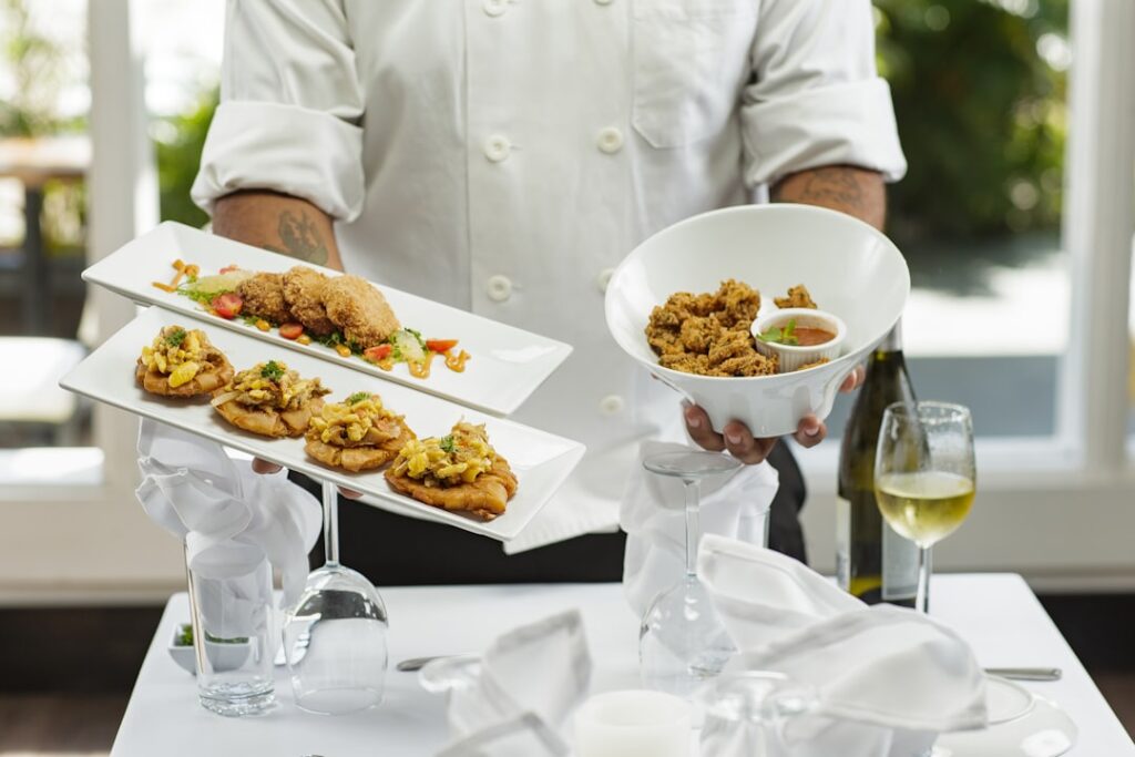 Chef in a white uniform presents two rectangular plates of bite-sized appetizers and a white bowl of fried chicken, on a white-tablecloth setup with a wine glass and bottle in the background.
